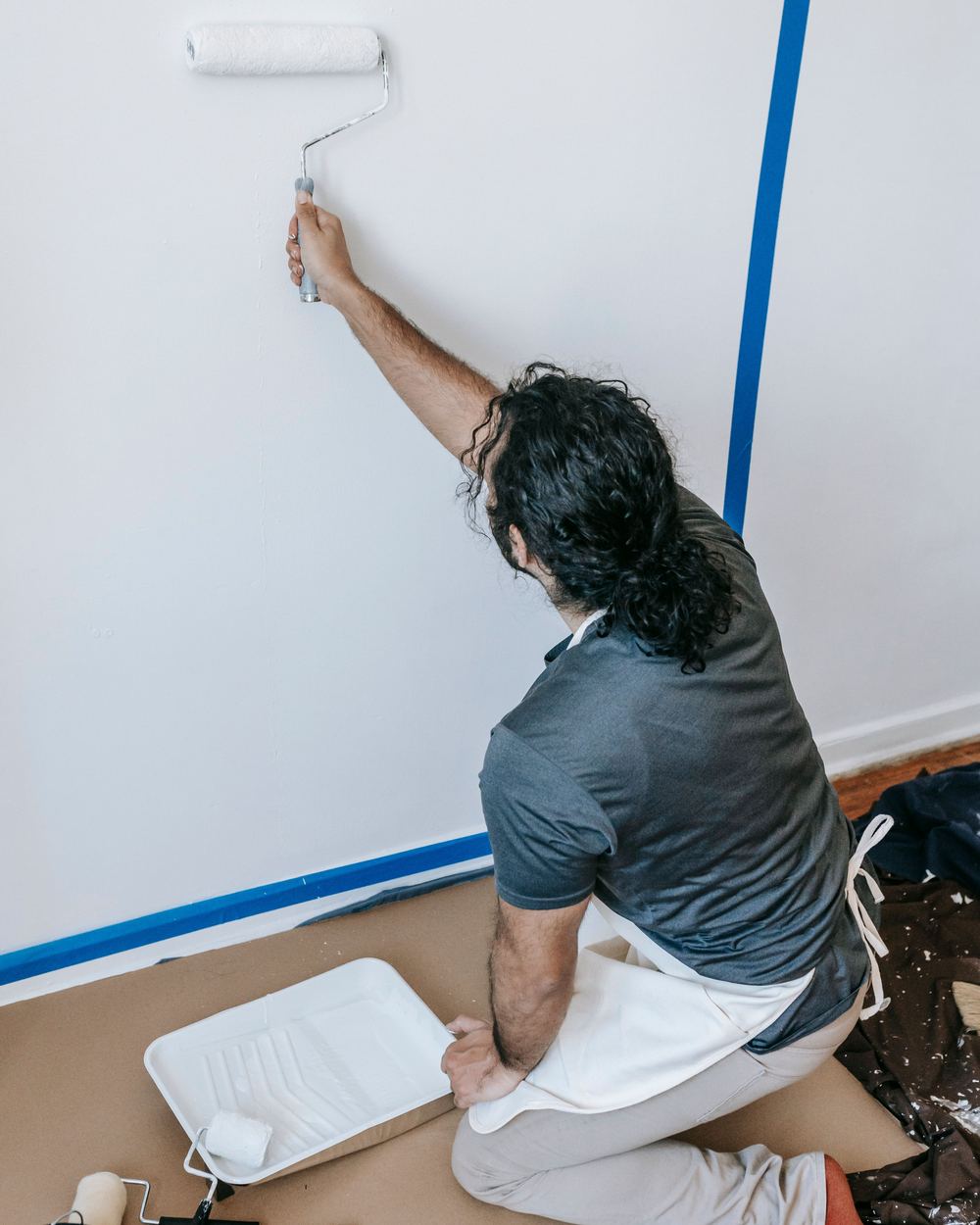 Een schilder rolt een witte muur, met afplaktape langs de randen en een verfbak op afdekpapier