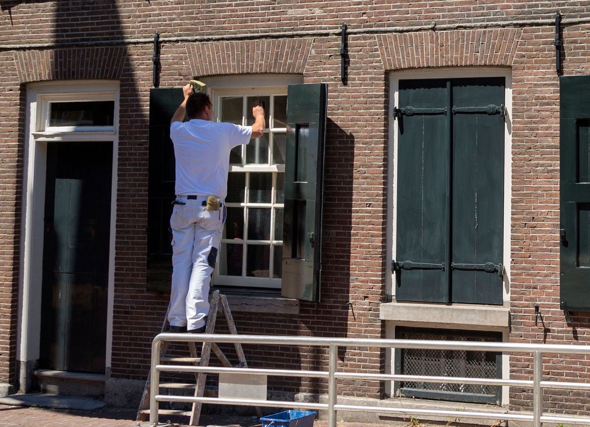 Een schilder op een ladder werkt een raam met groene luiken bij in een bakstenen gevel